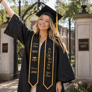 Può includere: Una toga e un tocco neri con nappa dorata, indossati con una stola di laurea nera e oro. La stola presenta scritte dorate che riportano School Name, BSN, RN, Class of 2026, NURSING e Your Name, insieme a icone di caduceo e stetoscopio.