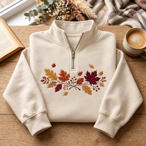 Könnte beinhalten: Ein cremefarbenes Sweatshirt mit kurzem Reißverschluss am Kragen, verziert mit einer Stickerei aus Herbstblättern in Gelb-, Orange-, Rot- und Dunkelburgundertönen sowie kleinen braunen Beeren, die mittig auf der Brust platziert ist.