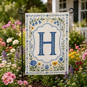 May include: A garden flag featuring a large blue letter "H" on a cream background, surrounded by a floral border with blue, yellow, and white flowers. The flag is displayed on a black metal stand in a garden setting.