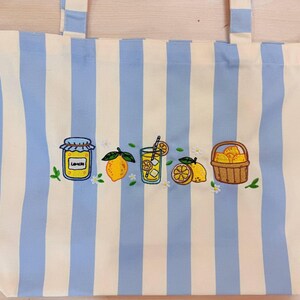 May include: A tote bag featuring vertical light blue and white stripes with an embroidered design across the center. The embroidery depicts a jar labeled LEMON, a whole lemon, a glass of lemonade with a straw, lemon slices, and a basket of lemons.