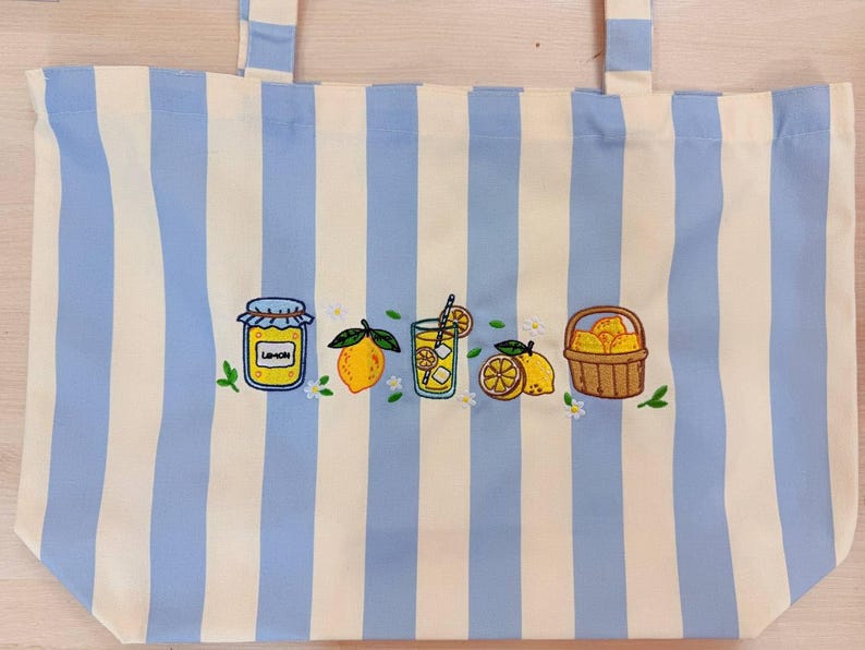 May include: A tote bag featuring vertical light blue and white stripes with an embroidered design across the center. The embroidery depicts a jar labeled LEMON, a whole lemon, a glass of lemonade with a straw, lemon slices, and a basket of lemons.