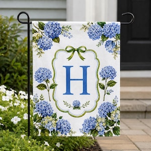 May include: A white garden flag with a blue letter "H" in a decorative frame, encircled by blue hydrangea flowers and green details. A green bow adorns the frame's top. The flag is displayed on a black metal stand.