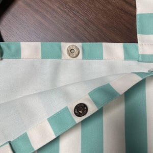 May include: A tote bag featuring a vertical striped pattern in mint green and white. The bag includes two wide fabric straps and a silver-toned magnetic snap closure located at the center of the top opening to secure the contents of the bag.