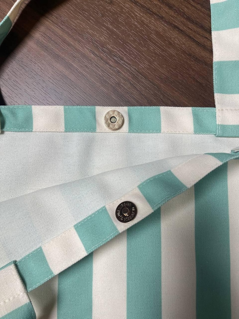 May include: A tote bag featuring a vertical striped pattern in mint green and white. The bag includes two wide fabric straps and a silver-toned magnetic snap closure located at the center of the top opening to secure the contents of the bag.