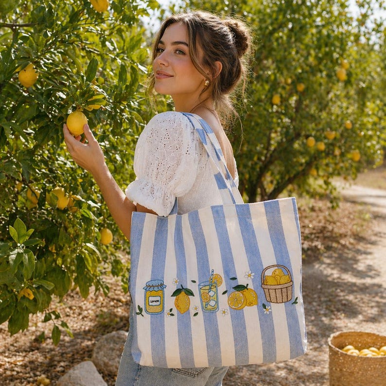 May include: A light blue and white vertical striped tote bag featuring embroidered illustrations of a jar of preserves, a whole lemon, a glass of lemonade with a straw, and a basket filled with lemons, accented with small white flower motifs.