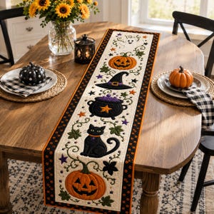 Puede incluir: Un camino de mesa acolchado rectangular con motivos bordados de Halloween que incluyen calabazas naranjas, un gato negro, un caldero morado, un sombrero de bruja y un fantasma, unidos por enredaderas verdes sobre un fondo crema con borde negro.