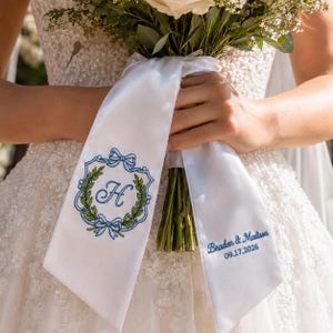 Op de afbeelding: Een wit satijnen lint gebonden rond een boeket bloemen. Het lint heeft blauwe geborduurde details, waaronder een monogram "H" in een krans en de namen "Braden & Madison" met de datum 09.17.2026.