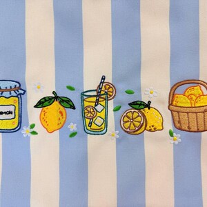 May include: A fabric featuring vertical light blue and cream stripes embroidered with a series of lemon-themed motifs including a jar labeled Lemon, a whole lemon, a glass of lemonade with a straw, sliced lemons, and a basket filled with lemons.