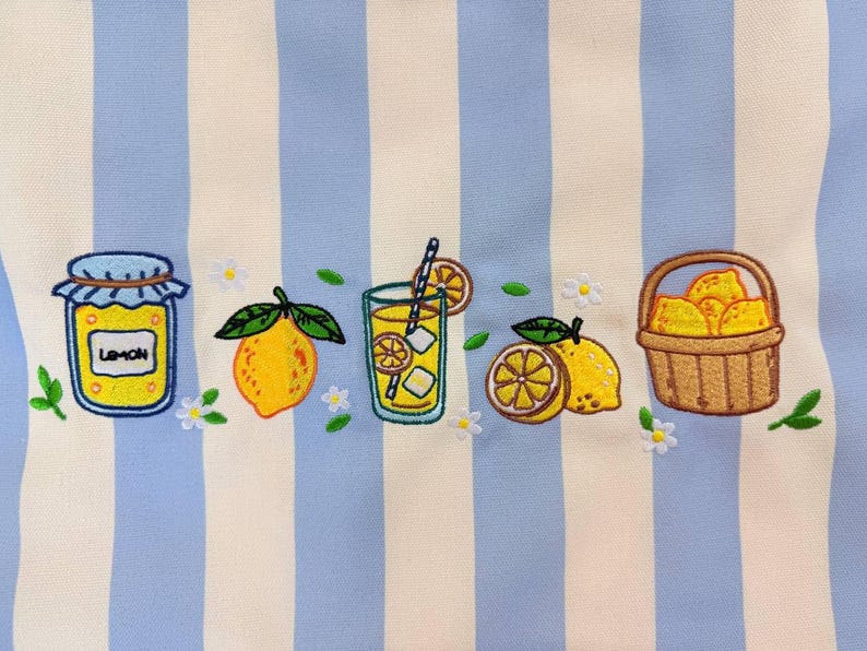May include: A fabric featuring vertical light blue and cream stripes embroidered with a series of lemon-themed motifs including a jar labeled Lemon, a whole lemon, a glass of lemonade with a straw, sliced lemons, and a basket filled with lemons.