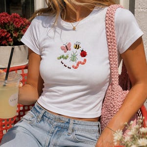 May include: A white short-sleeved crew neck t-shirt featuring a central embroidered design of various colourful insects, including a pink butterfly, a yellow and black bee, a red ladybird, a green caterpillar, a light green beetle, and a pink worm.
