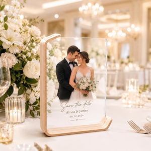 Pode incluir: Uma placa retangular de acrílico transparente numa base de madeira clara, com uma fotografia impressa de um casal, o texto "Save the date", os nomes "Alissa & James" e a data "08.30.2026" com um pequeno ícone de coração abaixo.