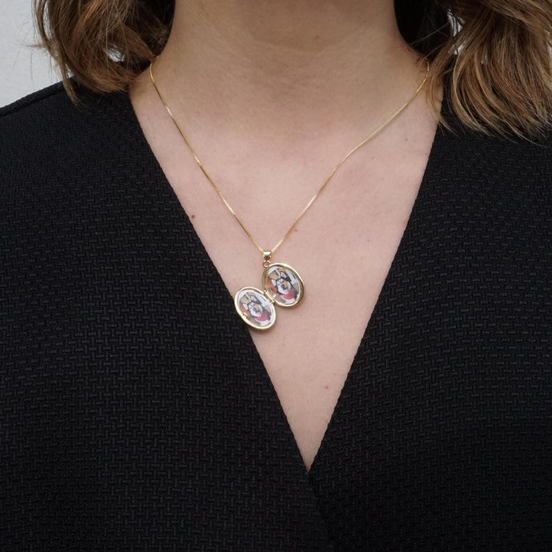 K&ouml;nnte beinhalten: Eine goldfarbene Halskette mit einer feinen Kette und einem kleinen, ovalen Medaillon. Das Medaillon, das sich &ouml;ffnet, um zwei kleine Bilder zu zeigen, wird vor einem schwarzen Kleidungsst&uuml;ck getragen.