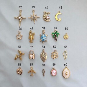 May include: A collection of gold-toned metal charms labeled 42 through 60, featuring various shapes including stars, a moon, a turtle, a palm tree, a lobster, a balloon dog, a padlock, a leaf, a clover, a wing, footprints, an airplane, and a cameo.