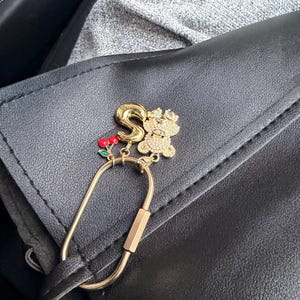 May include: A gold-colored keychain charm featuring the letter "S", a bejeweled teddy bear, and a cluster of red cherries. The charm is attached to a gold-colored carabiner clip, set against a black leather surface.