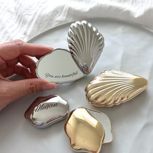 May include: A collection of shell-shaped compact mirrors in gold and silver metallic finishes. One open silver mirror displays the text 'You are beautiful', while a closed silver mirror is engraved with the name 'Megan'. Two other mirrors are closed.