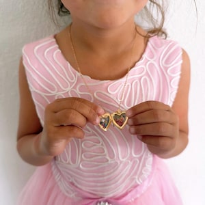 Op de afbeelding: Een gouden hartvormige medaillonketting met een ketting. Het medaillon is open en onthult twee kleine foto's. De ketting wordt vastgehouden door een persoon die een roze jurk draagt.