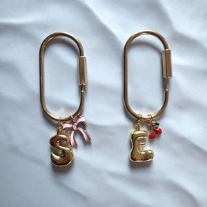 May include: Two gold-colored keychains with a carabiner-style clasp. One keychain has a gold letter "S" charm and a pink bow. The other has a gold letter "E" charm and a cherry charm.