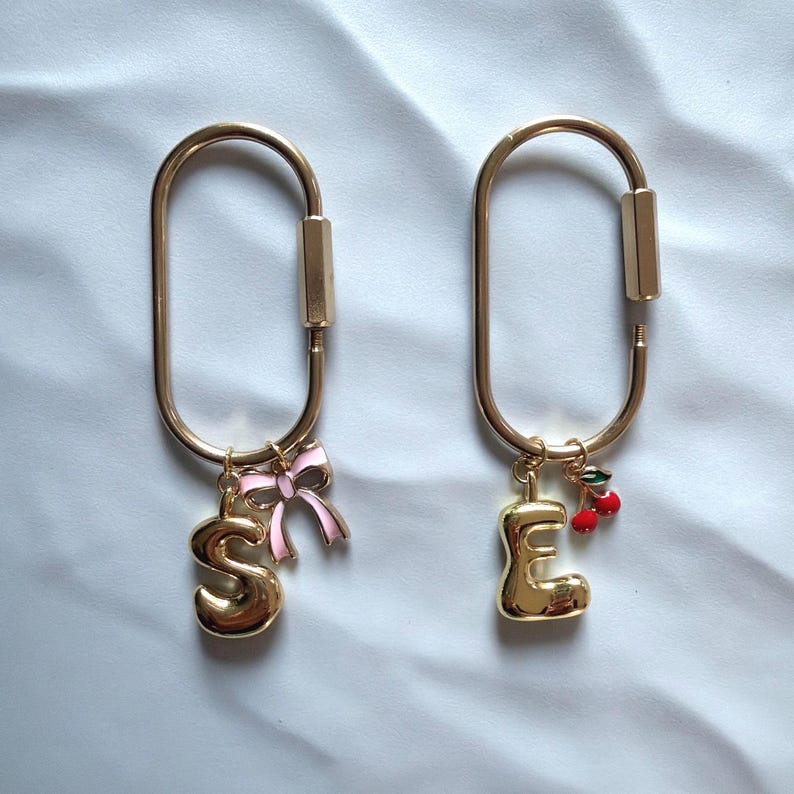 May include: Two gold-colored keychains with a carabiner-style clasp. One keychain has a gold letter "S" charm and a pink bow. The other has a gold letter "E" charm and a cherry charm.