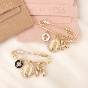 May include: Two gold-tone safety pins featuring dangling charms. One pin has a white cross, an oval Virgin Mary pendant, and a gold letter A. The other pin has a black cross, an oval Virgin Mary pendant, and a gold letter M. Both pins feature small white faux pearls.