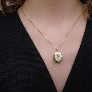 K&ouml;nnte beinhalten: Ein goldfarbener ovaler Medaillon-Anh&auml;nger mit Kreuzdesign, der an einer zarten Goldkette h&auml;ngt. Das Medaillon wird um den Hals getragen, vor einem dunklen Hintergrund.