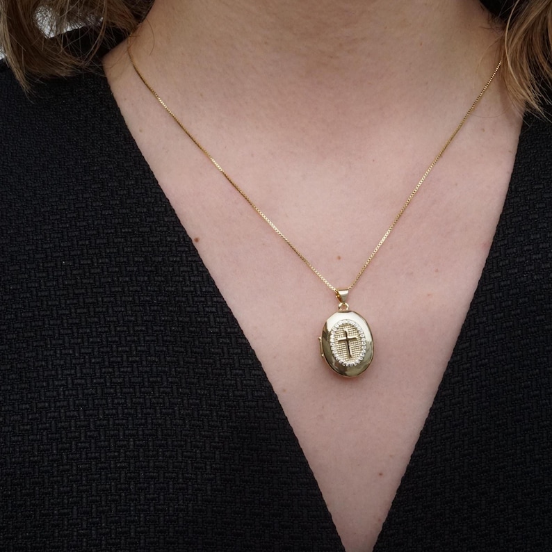 K&ouml;nnte beinhalten: Ein goldfarbener ovaler Medaillon-Anh&auml;nger mit Kreuzdesign, der an einer zarten Goldkette h&auml;ngt. Das Medaillon wird um den Hals getragen, vor einem dunklen Hintergrund.
