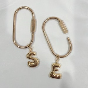 May include: Two gold-toned metal oval carabiner-style keychains, each featuring a polished, puffy letter charm. One keychain displays the letter S, while the other displays the letter E. Both carabiners have a screw-lock mechanism on the side.