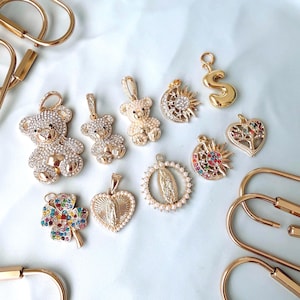 May include: A collection of gold-toned charms and pendants, including teddy bears, a crescent moon with a sun, a four-leaf clover, and a heart-shaped pendant. Some charms are embellished with crystals and colorful gemstones.