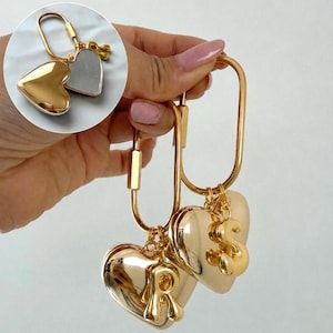 May include: Two gold-toned oval carabiner keychains, each featuring a smooth, polished heart-shaped charm. One keychain displays the letter R and the other displays the letter S. The inset shows the back of a heart charm with a silver-toned mirror finish.