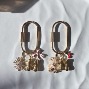 May include: Two gold-colored keychains with charms. One keychain has a sun charm, a gold letter "M", and a pink bow. The other keychain has a teddy bear charm, a gold letter "M", and a cherry charm. The keychains are on a white surface.