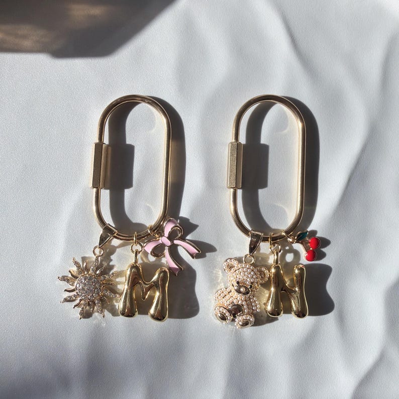 May include: Two gold-colored keychains with charms. One keychain has a sun charm, a gold letter "M", and a pink bow. The other keychain has a teddy bear charm, a gold letter "M", and a cherry charm. The keychains are on a white surface.