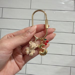 May include: Gold-toned keychain with a decorative charm arrangement. The charms include a pearl-studded teddy bear, a gold letter "S", and a cluster of red cherries. The keychain is held in a hand against a white tile background.