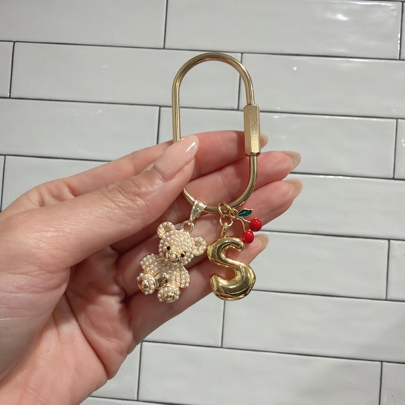 May include: Gold-toned keychain with a decorative charm arrangement. The charms include a pearl-studded teddy bear, a gold letter "S", and a cluster of red cherries. The keychain is held in a hand against a white tile background.