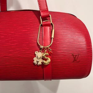 May include: A red leather handbag with a textured surface. The bag features a gold-colored charm with a teddy bear, the letter "S", and a cherry. The bag has a red leather strap and a gold-colored clasp.