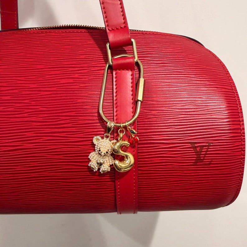 May include: A red leather handbag with a textured surface. The bag features a gold-colored charm with a teddy bear, the letter "S", and a cherry. The bag has a red leather strap and a gold-colored clasp.