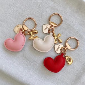 May include: Three keychains with heart-shaped charms in pink, white, and red. Each keychain has a gold-colored ring, a small heart charm with the word "LOVE", and a gold-colored letter or shape. The keychains are on a light gray surface.