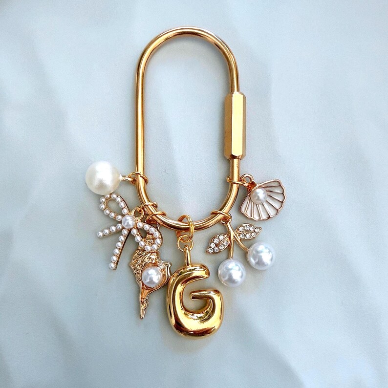 May include: A gold-toned oval carabiner keychain featuring a collection of charms including a letter G, a white pearl, a bow with small crystals, a seashell with a pearl, and a pair of cherries with crystal-encrusted leaves and pearl fruit.
