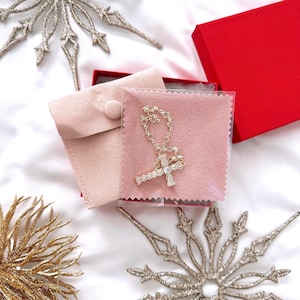 May include: A gold-colored necklace with a cross pendant, displayed on a pink cloth inside a clear plastic sleeve. The necklace is in a red box, with a pink pouch. Decorative silver and gold snowflake ornaments are in the background.