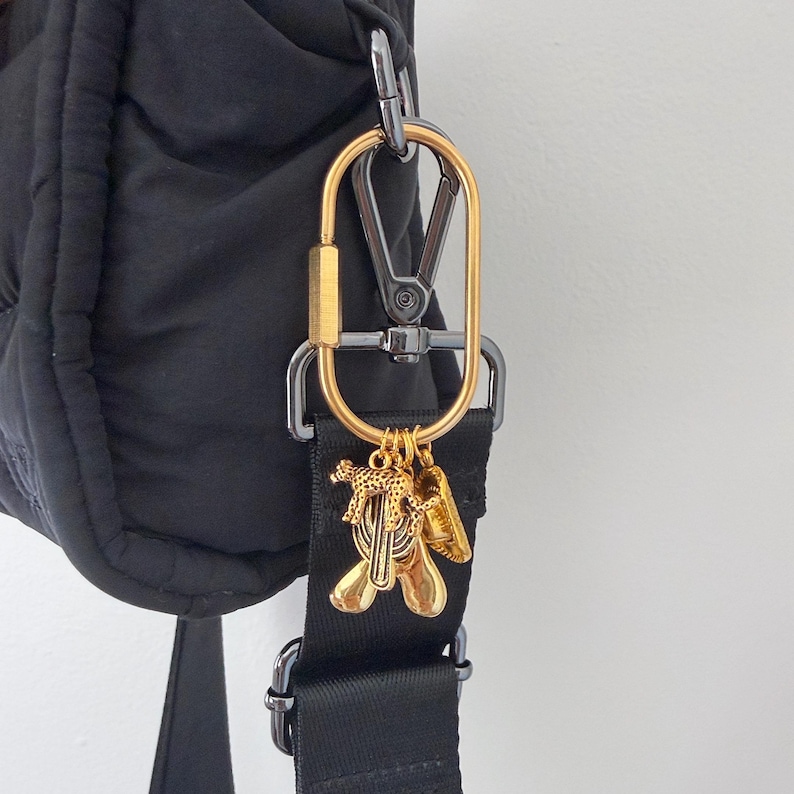 May include: A black quilted bag featuring a black nylon strap with a gold-toned oval carabiner clip. Attached to the carabiner are three small gold-toned charms, including a leopard figure, a circular disc, and a smooth, rounded abstract shape.