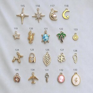 May include: A collection of gold-toned metal charms labeled 115 through 133, featuring various shapes including stars, a moon, a lobster, a turtle, a palm tree, a dog, a padlock, a leaf, a clover, a wing, footprints, an airplane, a key, and cameos.