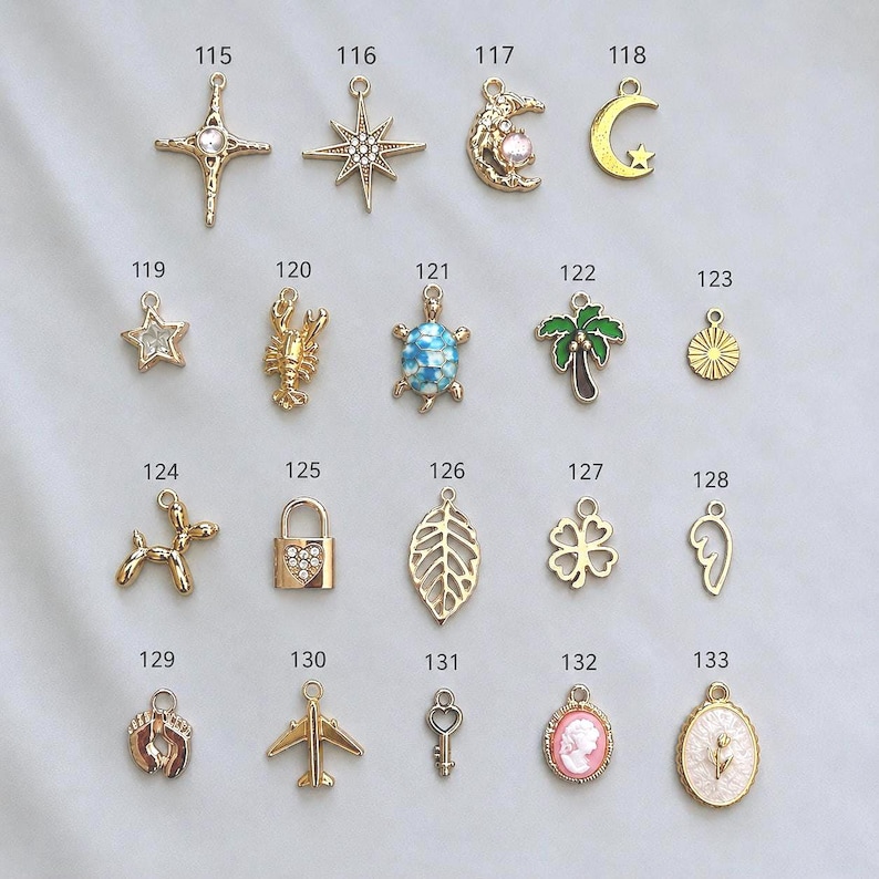 May include: A collection of gold-toned metal charms labeled 115 through 133, featuring various shapes including stars, a moon, a lobster, a turtle, a palm tree, a dog, a padlock, a leaf, a clover, a wing, footprints, an airplane, a key, and cameos.
