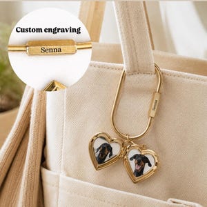 May include: A gold-toned metal carabiner keychain featuring two heart-shaped lockets that open to reveal photos of a black and tan dog. The carabiner has the custom engraving Senna on its side. The keychain is attached to a beige canvas bag.