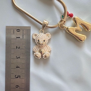 May include: A gold-colored keychain with a teddy bear pendant covered in small, pearl-like beads. The keychain also features a gold letter "M" charm and a small cluster of red cherries with green leaves.