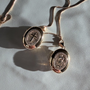 K&ouml;nnte beinhalten: Zwei goldfarbene ovale Medaillons an filigranen Ketten. Eines zeigt ein Kreuz, das andere eine religi&ouml;se Figur. Beide sind mit kleinen, funkelnden Steinen verziert. Der Schmuck liegt auf einer hellblauen Oberfl&auml;che.