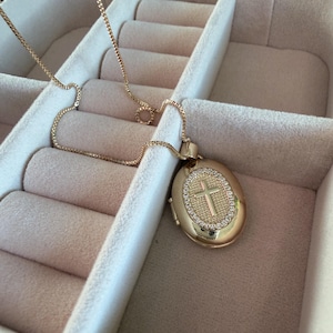 K&ouml;nnte beinhalten: Goldfarbene Halskette mit einem ovalen Anh&auml;nger mit Kreuzdesign. Der Anh&auml;nger ist mit kleinen, funkelnden Steinen um das Kreuz verziert. Die Halskette wird in einer Schmuckschatulle pr&auml;sentiert.