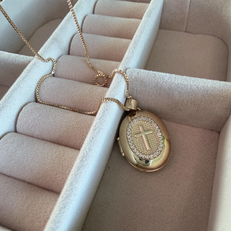 K&ouml;nnte beinhalten: Goldfarbene Halskette mit einem ovalen Anh&auml;nger mit Kreuzdesign. Der Anh&auml;nger ist mit kleinen, funkelnden Steinen um das Kreuz verziert. Die Halskette wird in einer Schmuckschatulle pr&auml;sentiert.