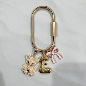 May include: Gold-toned keychain with a teddy bear charm covered in rhinestones, a gold letter "E" charm, and a pink bow charm. The keychain has a large oval shape with a clasp.