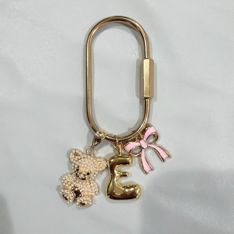 May include: Gold-toned keychain with a teddy bear charm covered in rhinestones, a gold letter "E" charm, and a pink bow charm. The keychain has a large oval shape with a clasp.