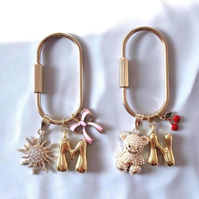 May include: Two gold-colored keychains with decorative charms. One keychain features a sun charm, the letter "M", and a pink bow. The other has a teddy bear, the letter "N", and a cherry charm.