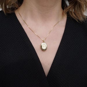 K&ouml;nnte beinhalten: Ein goldfarbener, ovaler Medaillon-Anh&auml;nger mit Kreuzmuster, der an einer feinen Goldkette h&auml;ngt. Das Medaillon wird um den Hals getragen, vor einem dunklen Hintergrund.