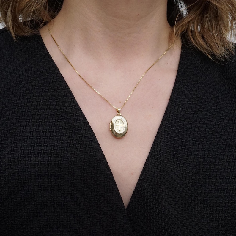 K&ouml;nnte beinhalten: Ein goldfarbener, ovaler Medaillon-Anh&auml;nger mit Kreuzmuster, der an einer feinen Goldkette h&auml;ngt. Das Medaillon wird um den Hals getragen, vor einem dunklen Hintergrund.